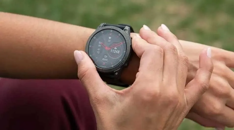 Close-up of a grey smartwatch on a wrist, displaying time and fitness data. Hands are adjusting the watch. Background is blurred green.