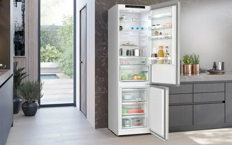 Réfrigérateur blanc ouvert, rempli de nourriture, dans une cuisine moderne avec vue sur un jardin.