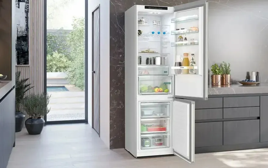 Réfrigérateur blanc ouvert, rempli de nourriture, dans une cuisine moderne avec vue sur un jardin.