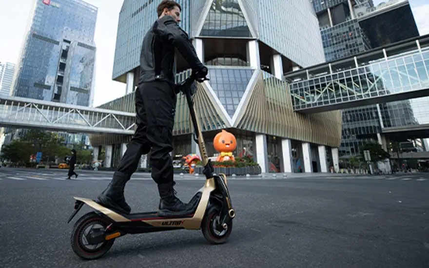 Een persoon in zwarte kleding rijdt op een elektrische step door een moderne zakenwijk met hoge glazen gebouwen en loopbruggen