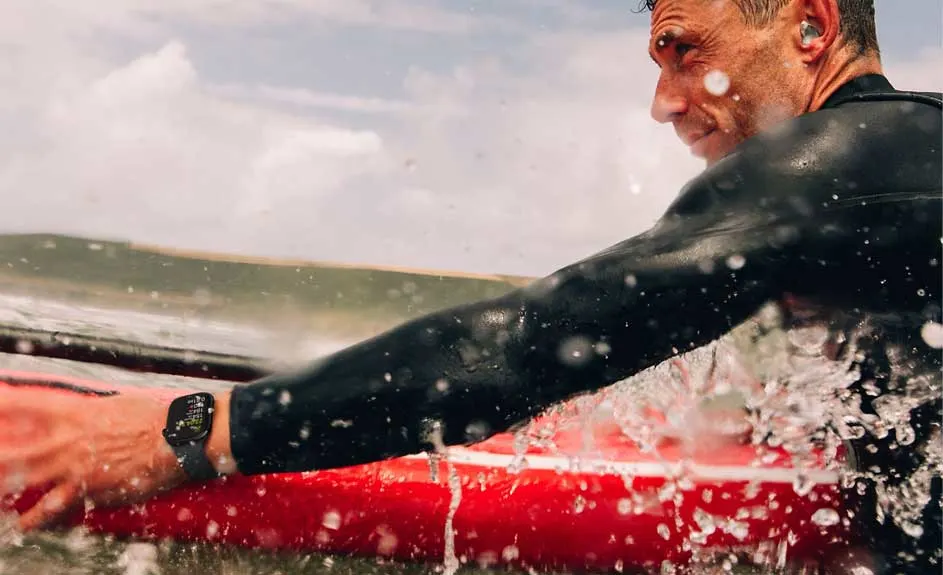 Un surfeur en combinaison noire sur une planche rouge dans l'eau, portant une montre. Ciel nuageux en arrière-plan.