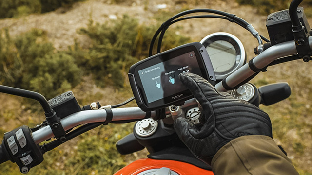 Gros plan sur une moto orange avec un GPS fixé au guidon gris. La main d'une personne interagit avec l'écran. Arrière-plan broussailleux.