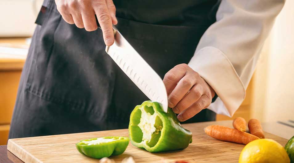 Un cuisinier coupe un poivron vert sur une planche à découper en bois. Des carottes et un citron sont à proximité.