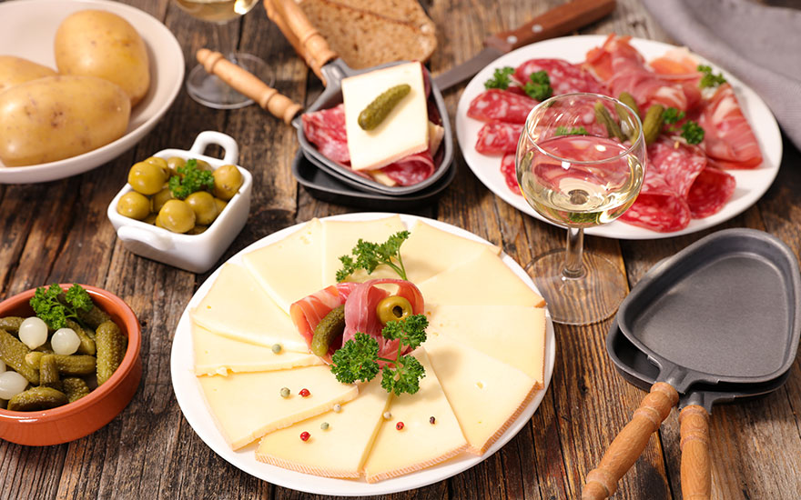Een houten tafel met raclette ingrediënten: kaas, vlees, augurken, zilveruitjes, olijven, aardappelen en witte wijn.
