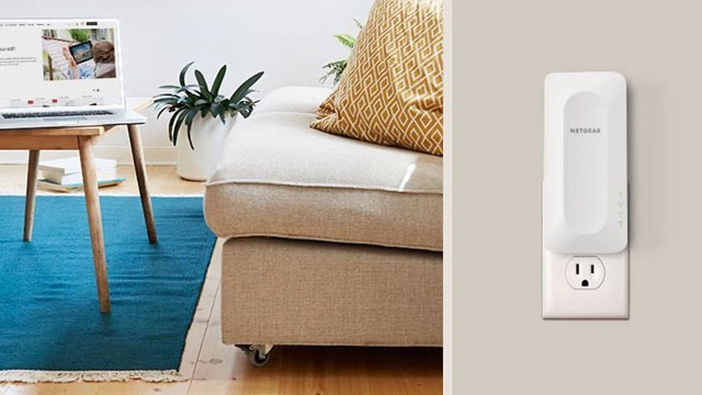 Een kamer met een laptop op een houten tafel, een witte bank met een geel kussen en een witte wifi-versterker in een stopcontact.