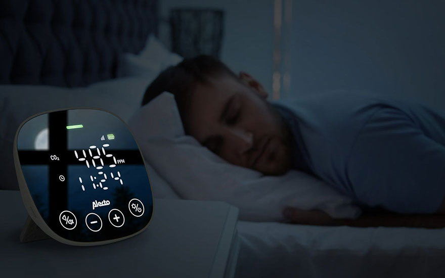 Een Alecto CO2-meter toont 485 ppm en 11:24 in een donkere slaapkamer, naast een slapende persoon op een wit kussen.