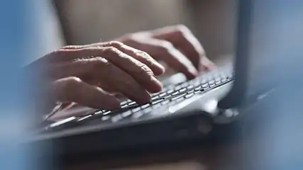 Mains tapant sur un clavier d'ordinateur portable gris. L'arrière-plan est flou, avec des tons neutres et une lumière douce.