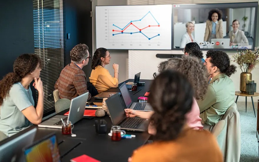 A large group of colleagues meets, looking at a graph on a big screen, while a video meeting with three people is ongoing.
