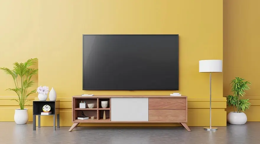 Un téléviseur noir est monté sur un meuble TV en bois devant un mur jaune. Une lampe et des plantes sont disposées sur le sol gris.