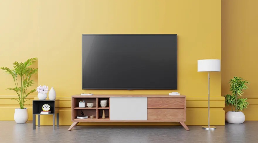 A living room scene with a large television above a wooden cabinet. Plants flank a lamp and a side table against a yellow wall.
