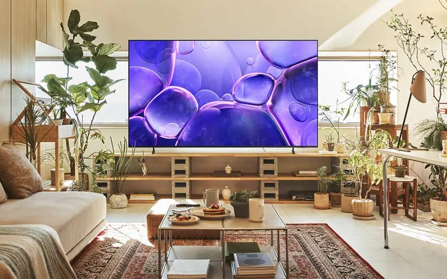 Een lichte woonkamer vol planten toont een grote tv met paarse abstracte vormen, omringd door houten meubels en zacht daglicht.