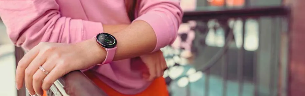 Een vrouw draagt een roze smartwatch terwijl ze buiten staat en haar pols controleert, met een moderne stedelijke omgeving op de achtergrond.
