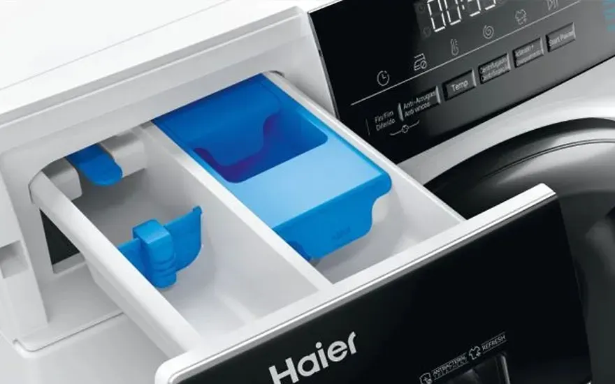 Een close-up van het wasmiddellade van een witte Haier wasmachine met blauwe vakjes en een digitaal display, modern en gebruiksvriendelijk.