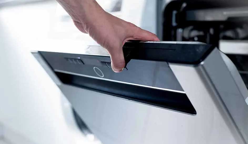 Une main ouvre la porte grise et noire d'un lave-vaisselle moderne dans une cuisine éclairée.