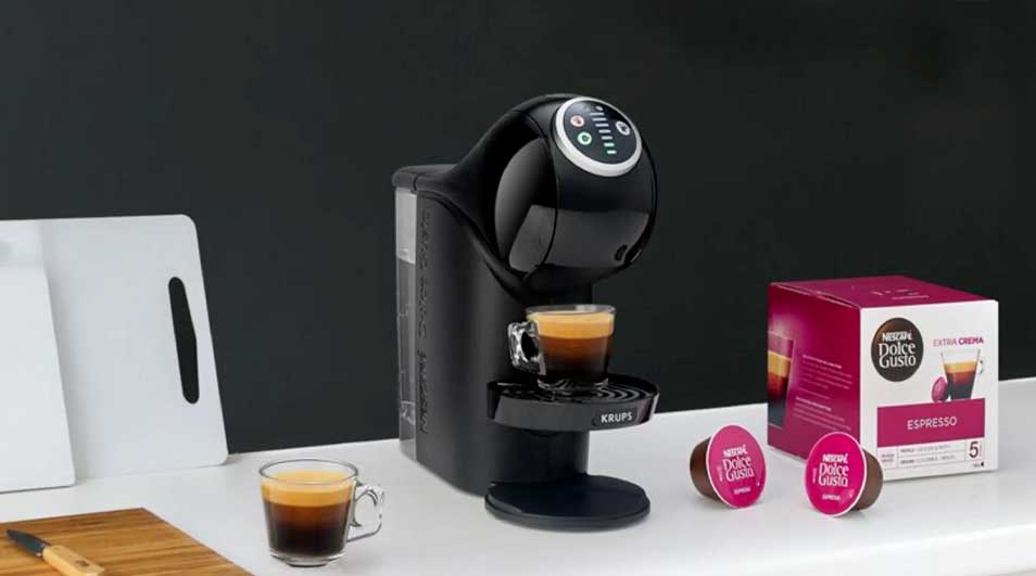 Une machine Dolce Gusto noire prépare un espresso sur un plan blanc, près de capsules roses, d'une boîte, de planches et d'un évier.