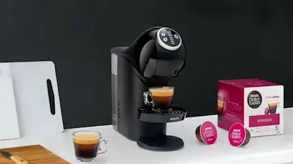 Une machine Dolce Gusto noire prépare un espresso sur un plan blanc, près de capsules roses, d'une boîte, de planches et d'un évier.