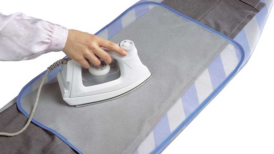 Une main repasse un pantalon gris sur une planche à repasser bleue et blanche avec un fer blanc.