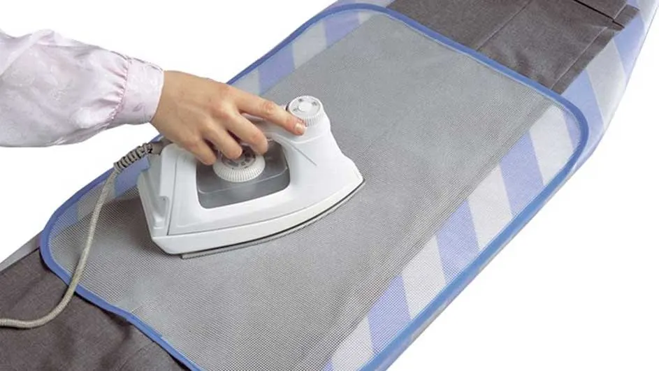 Une main repasse un pantalon gris sur une planche à repasser bleue et blanche avec un fer blanc.