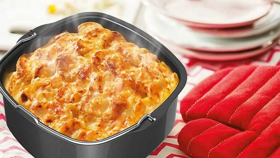 Ovenschotel in een zwarte bakvorm, naast rode ovenwanten op een rood-wit tafelkleed. Gestapelde witte borden op de achtergrond.