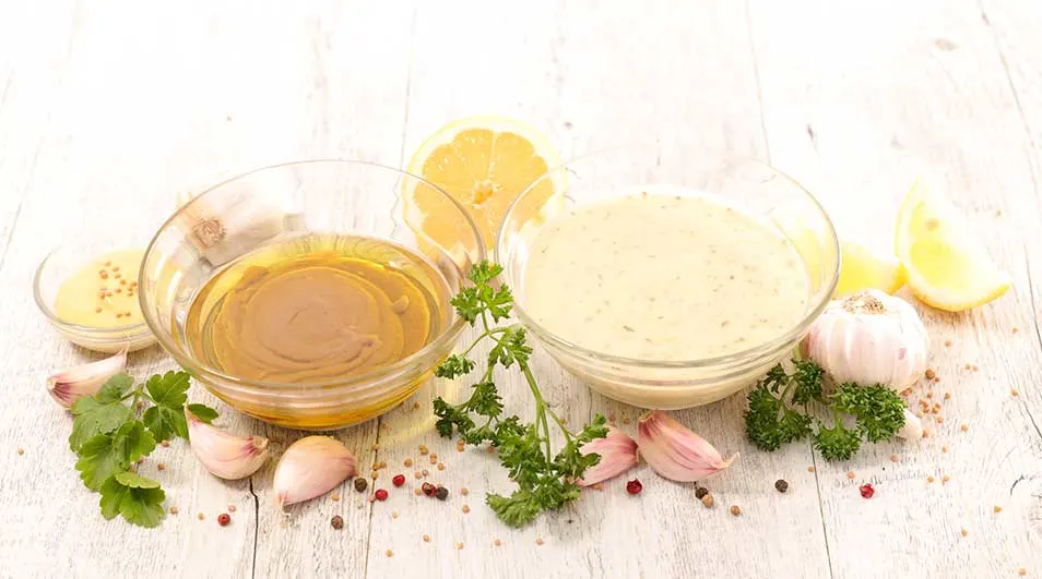 Twee glazen kommen met mosterd en mayonaise, citroenpartjes, knoflook en peterselie op een witte houten tafel.