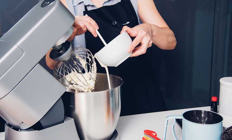 Une cuisine avec un robot culinaire gris, un bol en acier inoxydable et une tasse blanche versant du liquide.