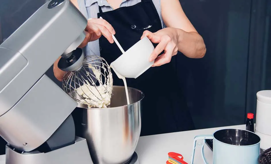 Une cuisine avec un robot culinaire gris, un bol en acier inoxydable et une tasse blanche versant du liquide.