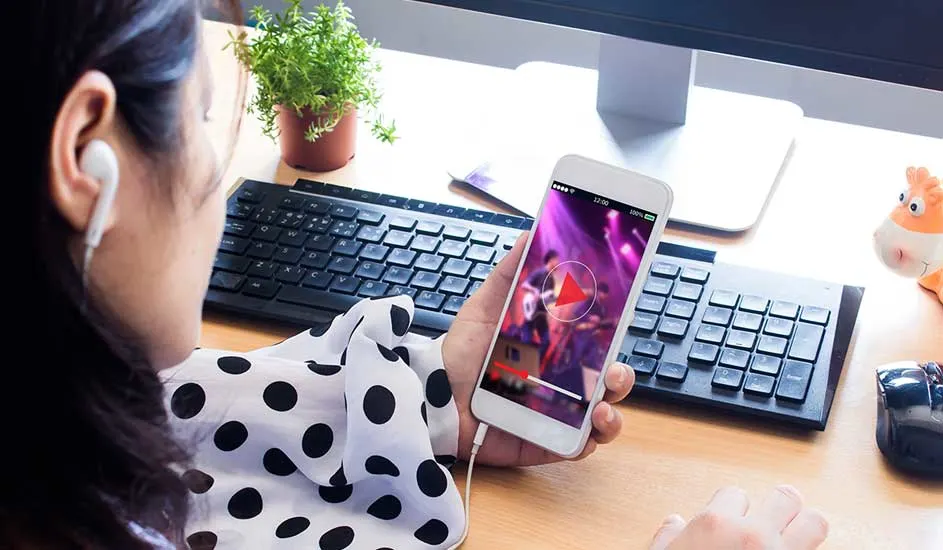 Une personne tient un smartphone avec une vidéo en lecture, entourée de claviers, d'un écran, d'une plante et d'une figurine.