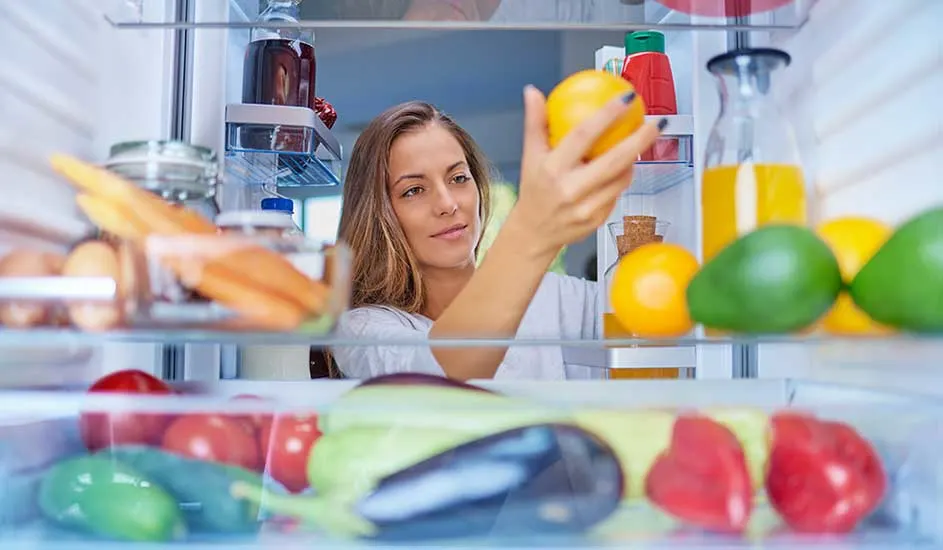 Frigo rempli de fruits et légumes, avec des oranges, des avocats, des tomates, des aubergines et des carottes visibles.