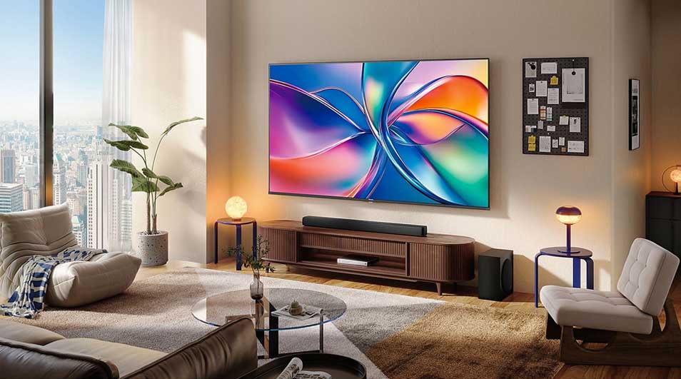 Un salon lumineux avec grande TV colorée, barre de son, meuble en bois, fauteuils, plantes et hautes fenêtres sur la ville.