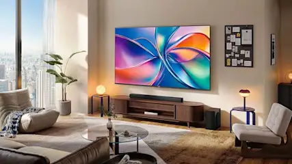 Un salon lumineux avec grande TV colorée, barre de son, meuble en bois, fauteuils, plantes et hautes fenêtres sur la ville.