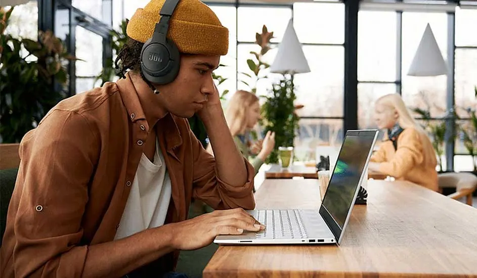 Een laptop op een houten tafel, met op de achtergrond ramen en planten. Iemand draagt een gele muts en een bruin hemd.
