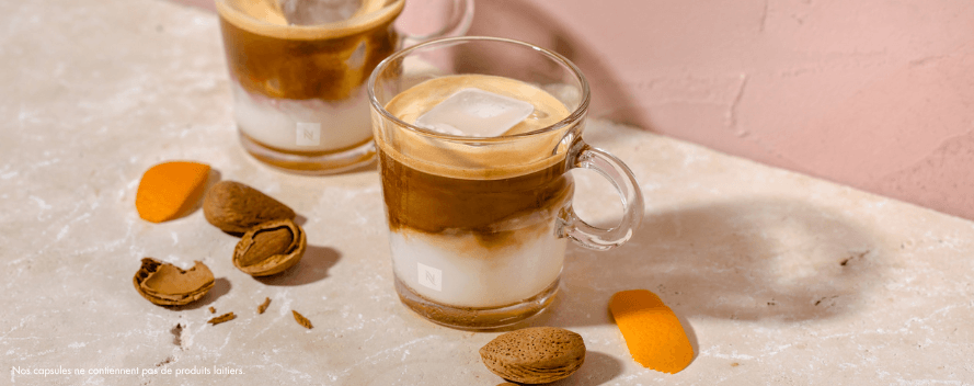 Deux verres de café glacé au lait, avec des amandes, des coquilles d’amandes brisées et des écorces d’orange sur une surface rose pâle. Une note en bas indique que les capsules ne contiennent pas d’ingrédients laitiers.