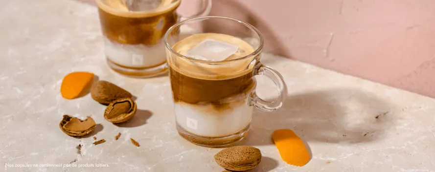 Deux verres de café glacé au lait, avec des amandes, des coquilles d’amandes brisées et des écorces d’orange sur une surface rose pâle. Une note en bas indique que les capsules ne contiennent pas d’ingrédients laitiers.