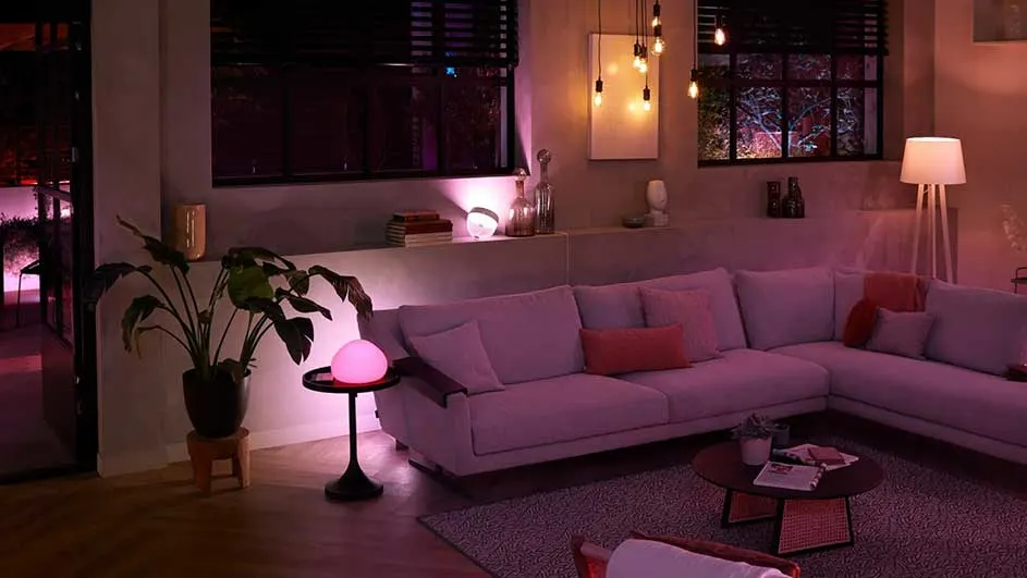 Een woonkamer met een grote hoekbank, planten, lampen en decoratieve objecten onder zacht, roze licht.
