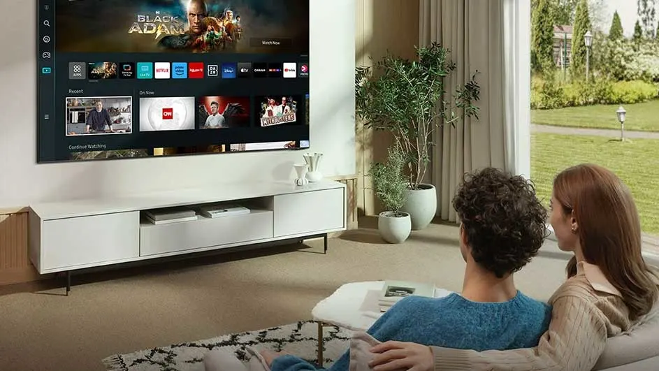 Woonkamer met tv aan de muur boven een wit tv-meubel. Er staan planten en een raam met uitzicht op een tuin.