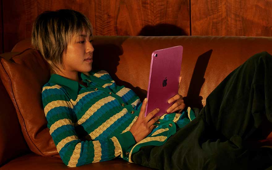 Une jeune personne se détend sur un canapé en cuir brun, tient un iPad rose et porte un cardigan rayé vert et crème sous une lumière chaude.