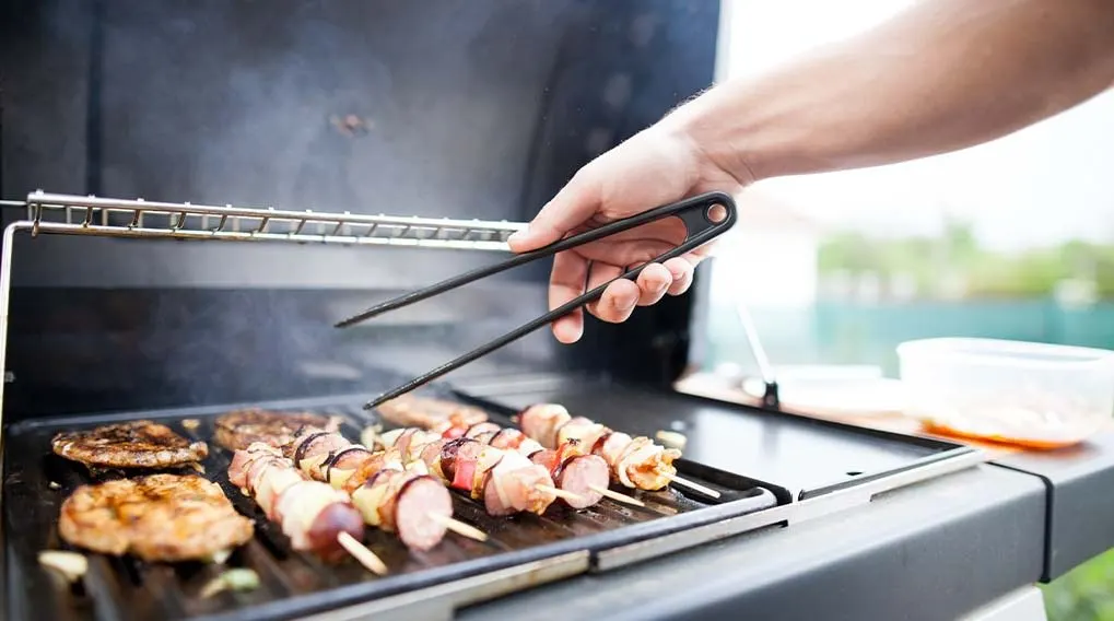 A grill with metal grates holds meat patties and skewers. A hand holds black tongs. Background is blurred with greenery.