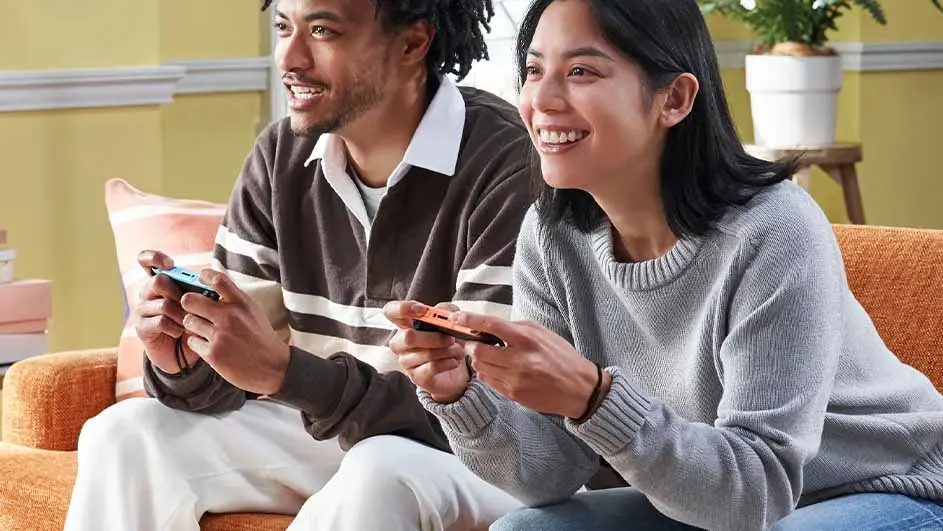 Twee personen op een oranje bank spelen spelletjes met blauwe en oranje controllers. Achtergrond: gele muur en plant.