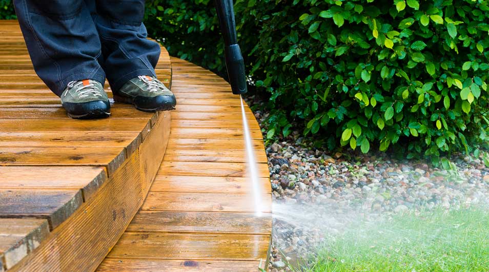 Een houten terras wordt gereinigd met een hogedrukreiniger, groene planten op de achtergrond.