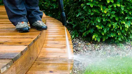Een houten terras wordt gereinigd met een hogedrukreiniger, groene planten op de achtergrond.