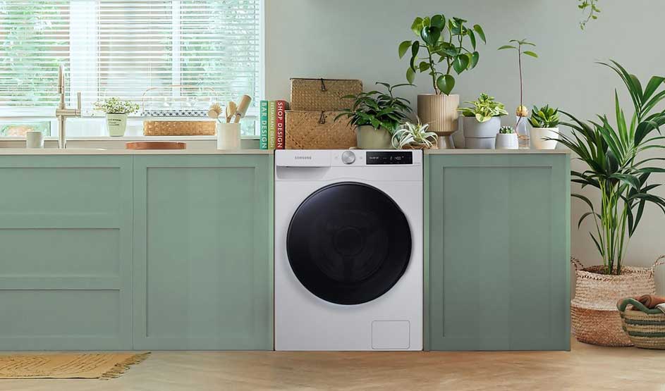 Een witte wasmachine staat ingebouwd tussen mintgroene kasten, omringd door planten en manden in een lichte, rustige ruimte.