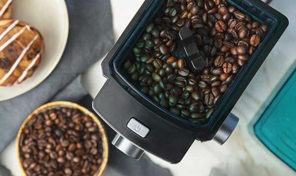 Een koffiemolen wordt van bovenaf gevuld met bruine bonen. Ernaast staan een kom met extra bonen en een bord met een gebakje, op een lichte tafel.