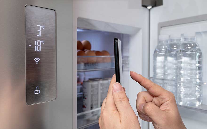 Een open koelkast met een digitaal display dat 3°C en -18°C toont, met flessen water en eieren zichtbaar binnenin. Een hand houdt een smartphone vast.