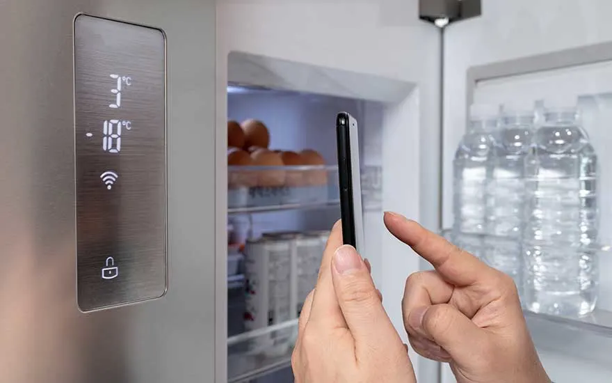 Een open koelkast met een digitaal display dat 3°C en -18°C toont, met flessen water en eieren zichtbaar binnenin. Een hand houdt een smartphone vast.