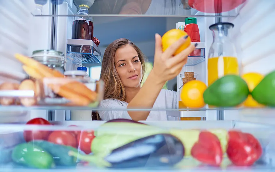 A refrigerator filled with food. Shelves display fruits, vegetables, and juice. An orange is being held.
