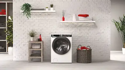 Wasmachine in een wasruimte met bakstenen muur, houten rek met handdoeken en een rieten wasmand met rode handdoeken.