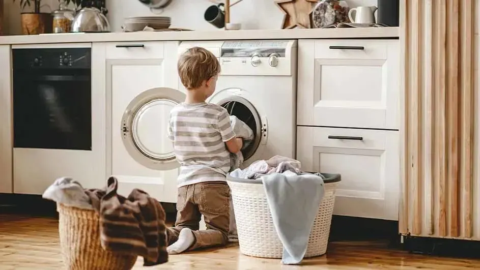 Een kind knielt voor een witte wasmachine en stopt wasgoed erin, met volle wasmanden ernaast, merk van de machine is niet zichtbaar.