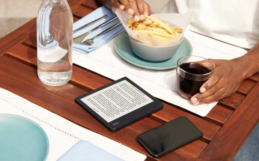 Een houten tafel met een fles water, frieten, een drankje, een e-reader en een telefoon.