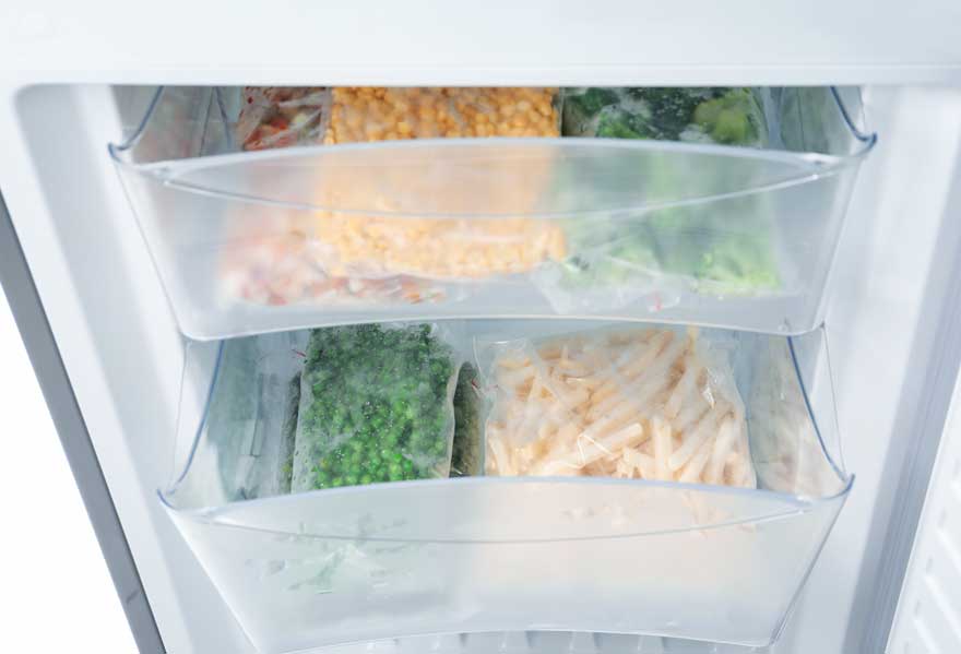 Een open witte diepvriezer met twee plastic bakken vol diepvriesproducten, zoals erwten, frieten, mais en broccoli.