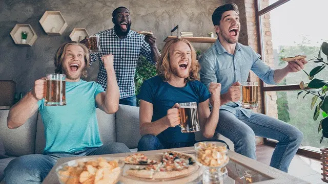 Quatre personnes regardent un match avec de la bière et des collations sur une table.